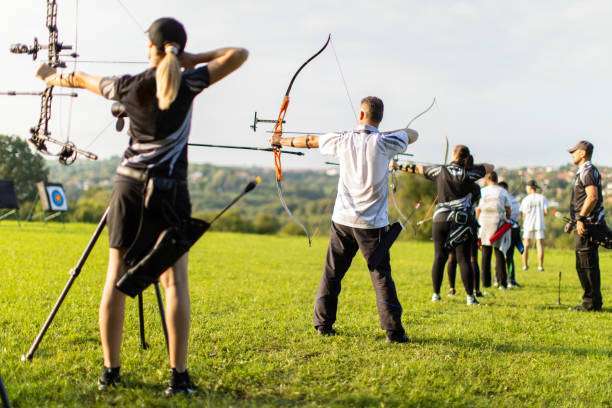 Women and men practicing archery training with recurve and compound bow on an open field before sunset.