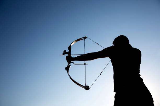 A silhouette of an archer draws his compound bow and aims upwards with a clean blue sky as a background.