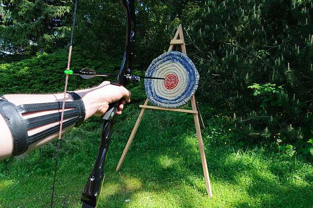 Archer aims a compound bow at an outdoor target with an arrow already hit.
