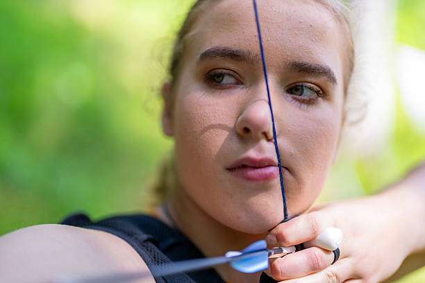 Young woman aims arrow with compound bow in forest setting.