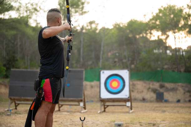 Man aims compound bow at target on outdoor archery range.