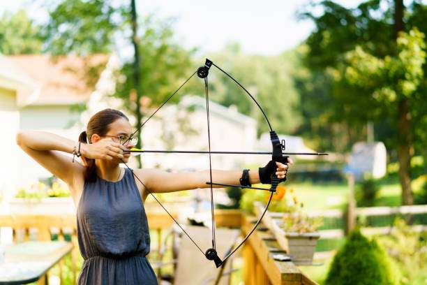 Woman with glasses aiming a black compound bow in a sunny backyard, practicing archery.