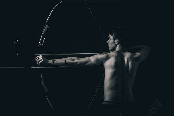Muscular man aiming a compound bow in a dark, dramatic black and white studio shot.