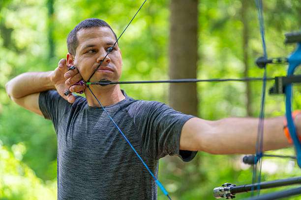 Man draws arrow with compound bow outdoors, focused on target.