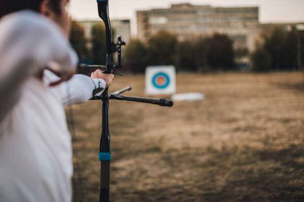 Person aiming a compound bow at an outdoor archery target in a field.