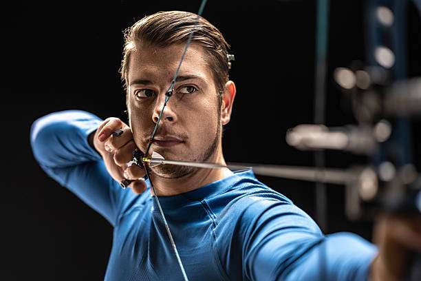 A male archer in a blue athletic shirt is captured in mid-draw, intently aiming his compound bow.
