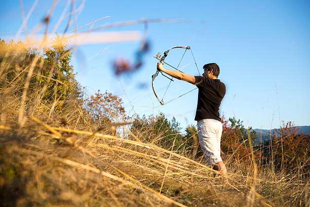 An archer drawing his compound bow in a field in the forest during the early autumn.
