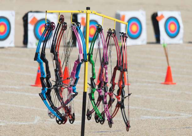 A man with a prosthetic arm practicing archery with a compound bow on the field in training at sunset. He is aiming directly at the camera. Focus on archer's face