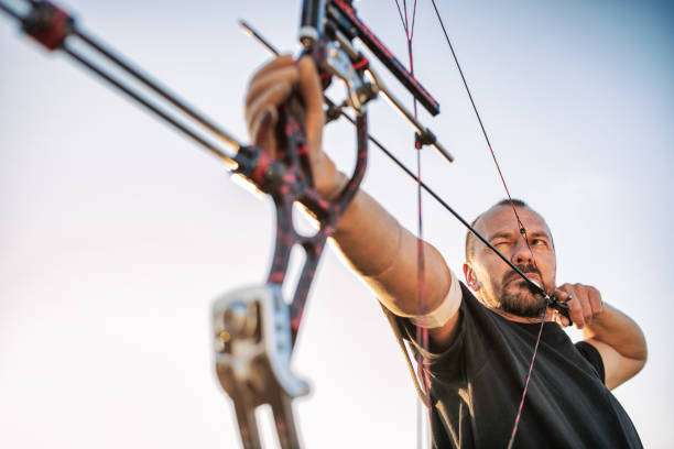 A man with a prosthetic arm practicing archery with a compound bow on the field in training at sunset. Focus on archer's face