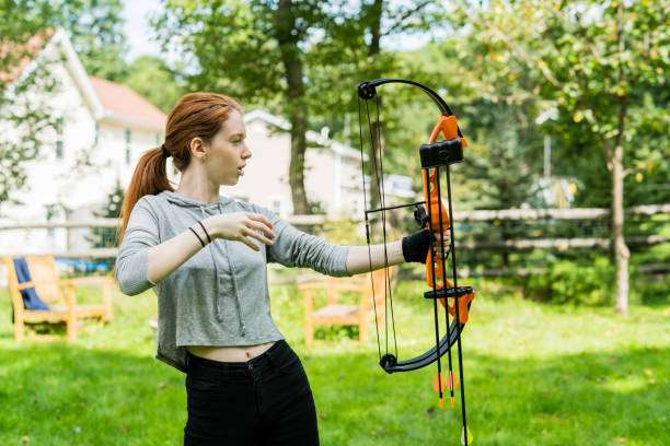 Young woman aiming an orange and black bow outdoors. Learning archery with a compound bow.