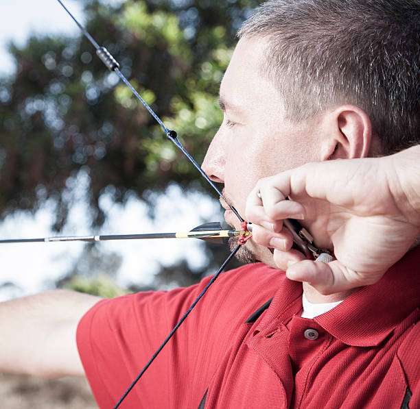 Focused male archer drawing a compound bow string, aiming his shot outdoors.