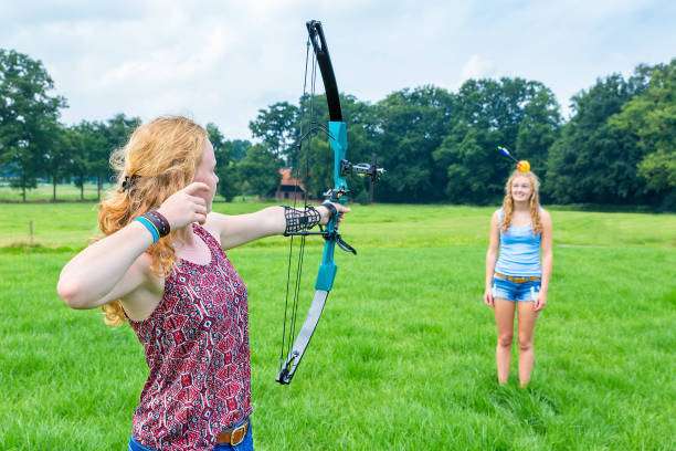 Dutch teenage girl aiming arrow of compound bow at fruit on head of young woman.