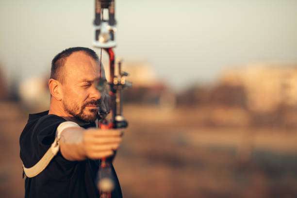 A man with a prosthetic arm practicing archery with a compound bow on the field in training at sunset. He is aiming directly at the camera. Focus on archer's face