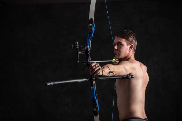 Muscular male archer in a strong stance with a compound bow on a dark background.