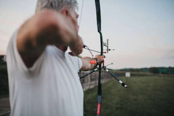 Rear view of an older man aiming a compound bow at a distant archery target.